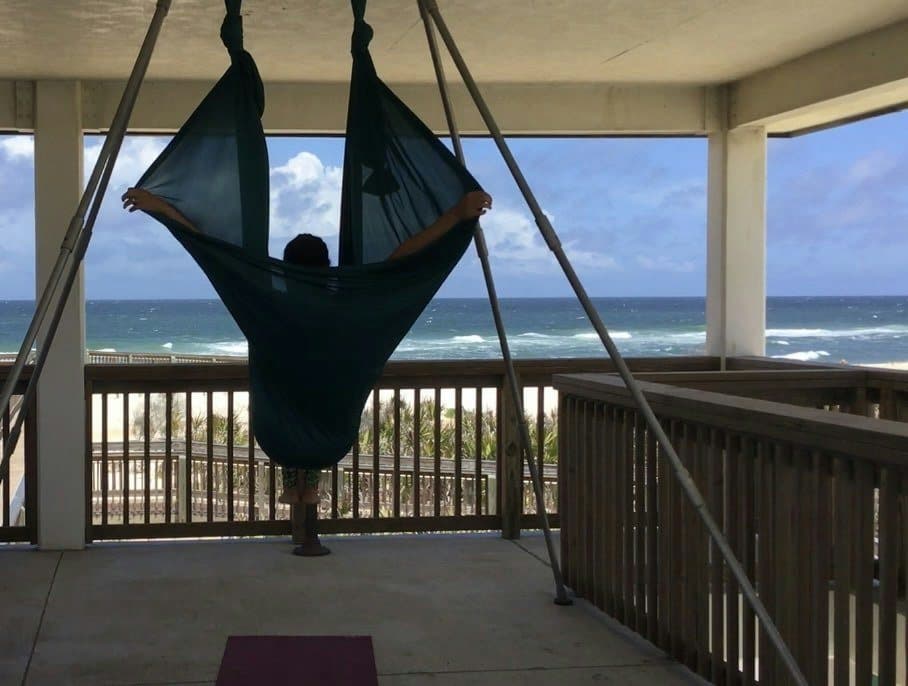 A student opens a teal hammock wide like wings, facing the ocean from the pavilion