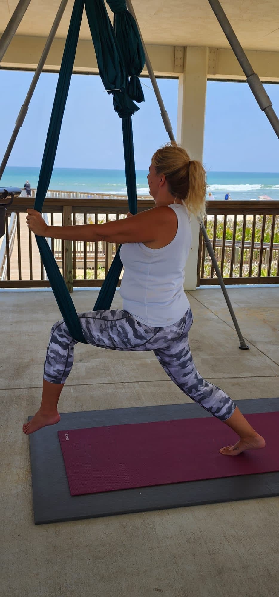 A student in a wide warrior stance holding a hammock at the beach pavilion, ocean behind her