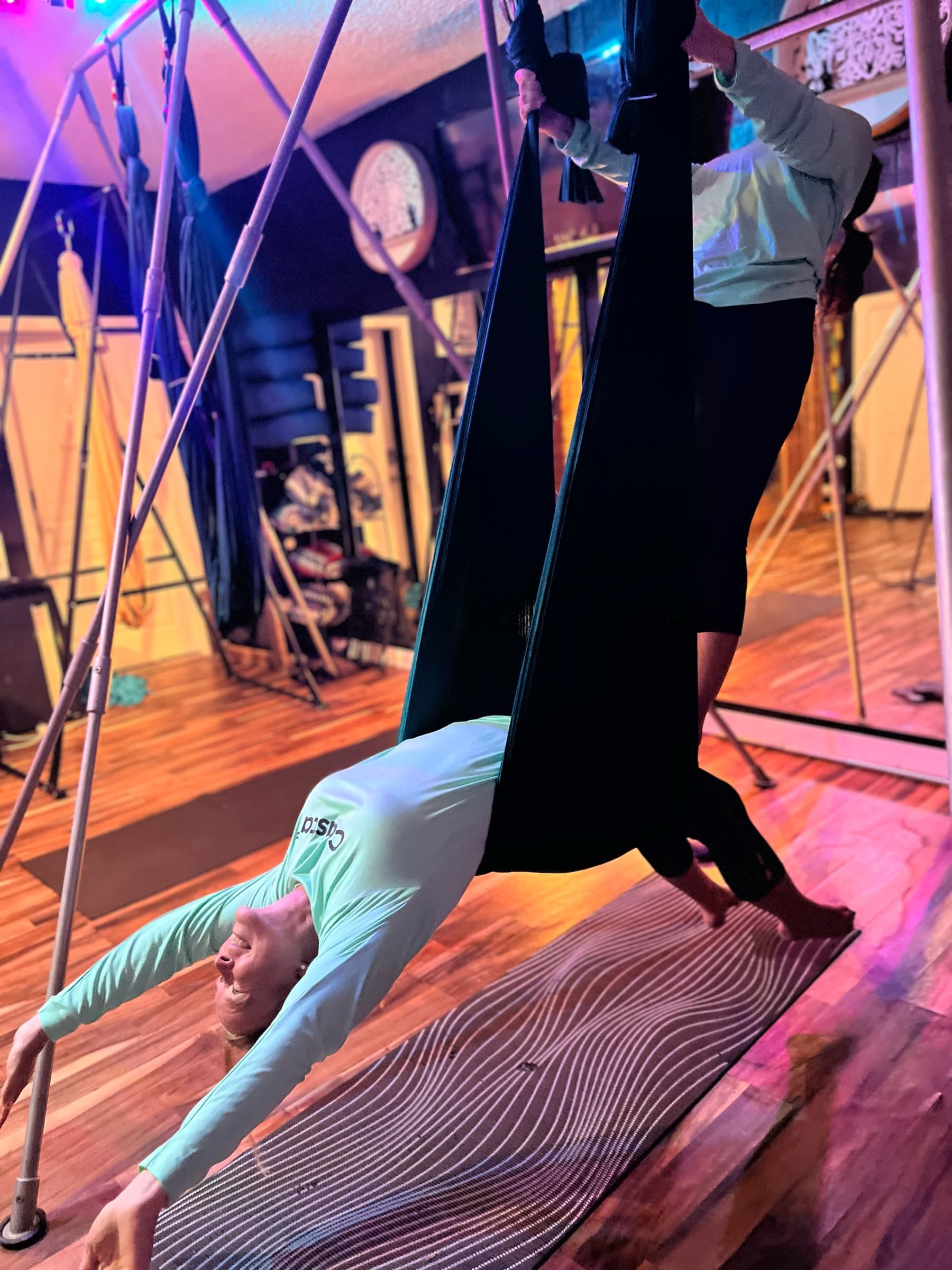 A student arches back through an aerial hammock inversion in the studio under pink and blue lights