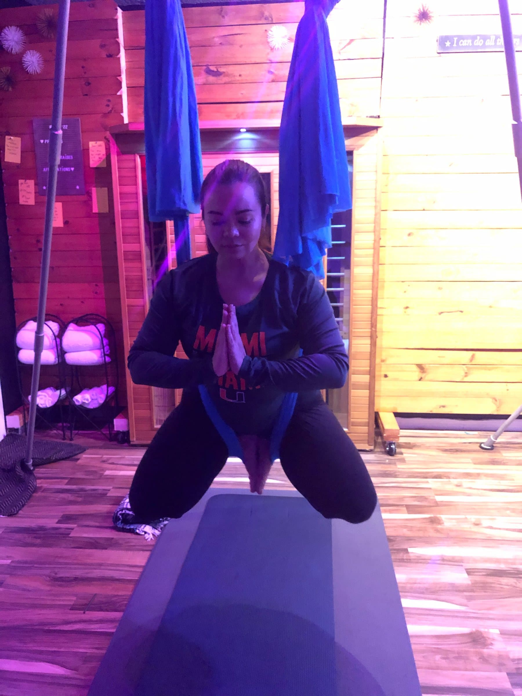 A student kneels in a blue hammock seat with hands at heart center, the studio glowing pink and gold
