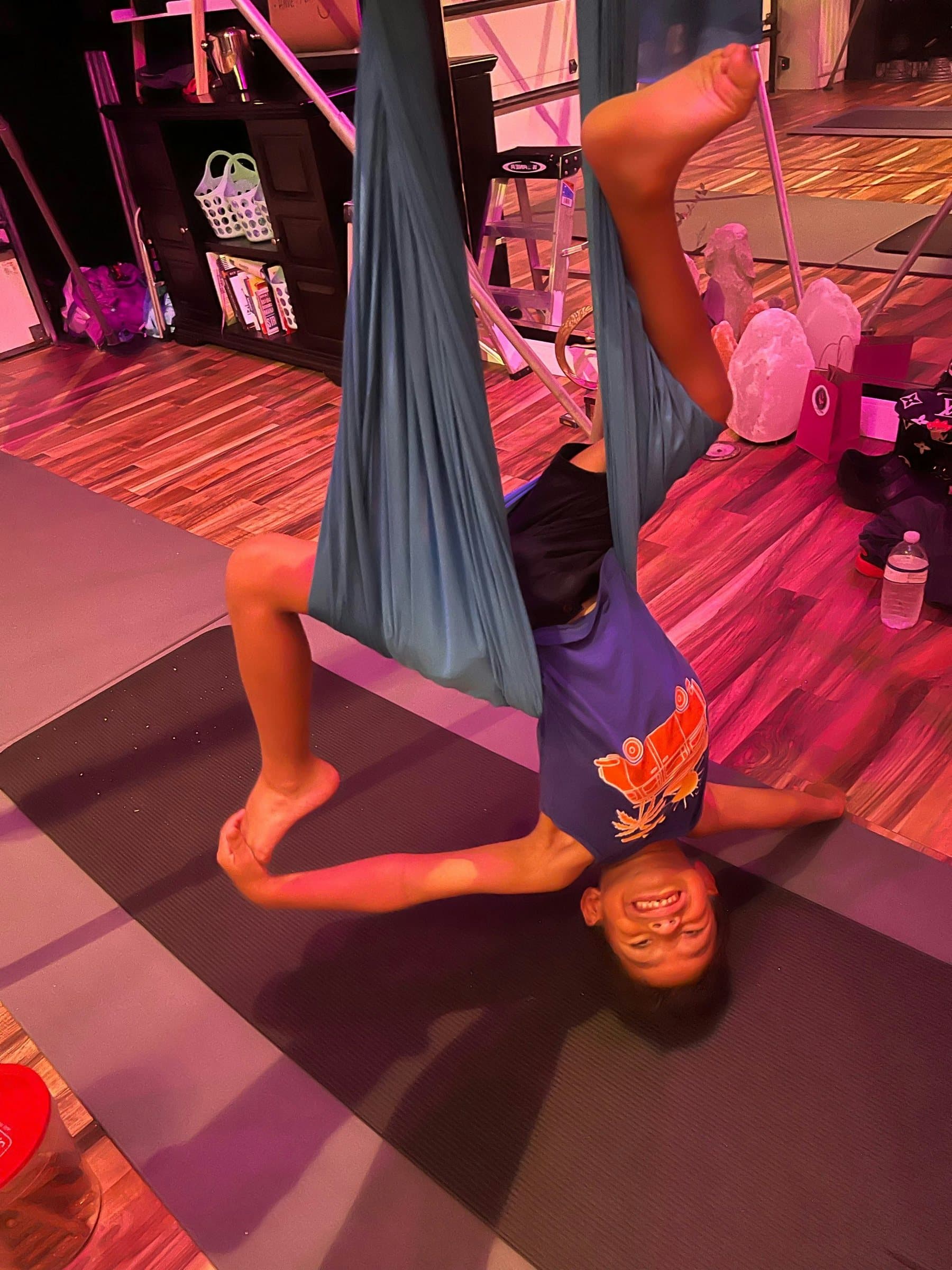 A young student hangs upside down in a teal hammock, grinning, in the studio