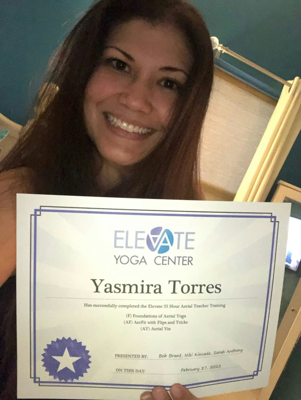 Yassy smiling and holding her Elevate Yoga Center aerial teacher training certificate
